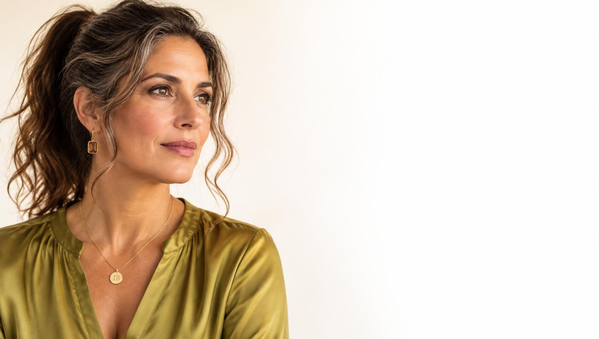 An experienced professional woman in a chartreuse silk blouse, wearing a gold pendant engraved TB, looking thoughtfully to the side.