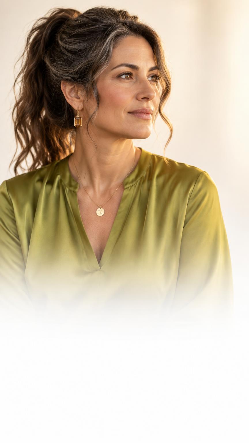 An experienced professional woman in a chartreuse silk blouse, wearing a gold pendant engraved TB, looking thoughtfully to the side.